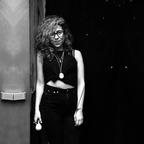 Ariana Reines standing in a doorway wearing black jeans, a black shirt, and several necklaces