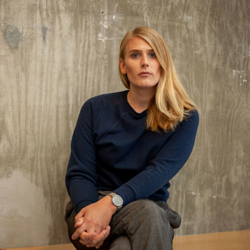 Jos Charles seated with her hands folded in her lap. She has shoulder length blonde hair and is wearing a navy blue sweater, gray pants, and a gray watch. She is in front of a gray wood background.