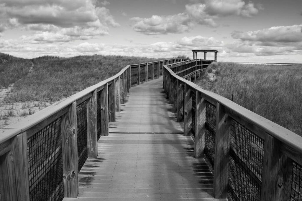 A black and white, digital photograph of a raised, wooden walkway through some sand dunes. The perspective of the photo has the viewer looking straight ahead on the wooden walkway. There are wooden railings on both sides of the walkway and in the distance, the path turns right and leads to a wooden gate. Patches of beach grass cover the dunes and the sky is full of cumulus clouds.
